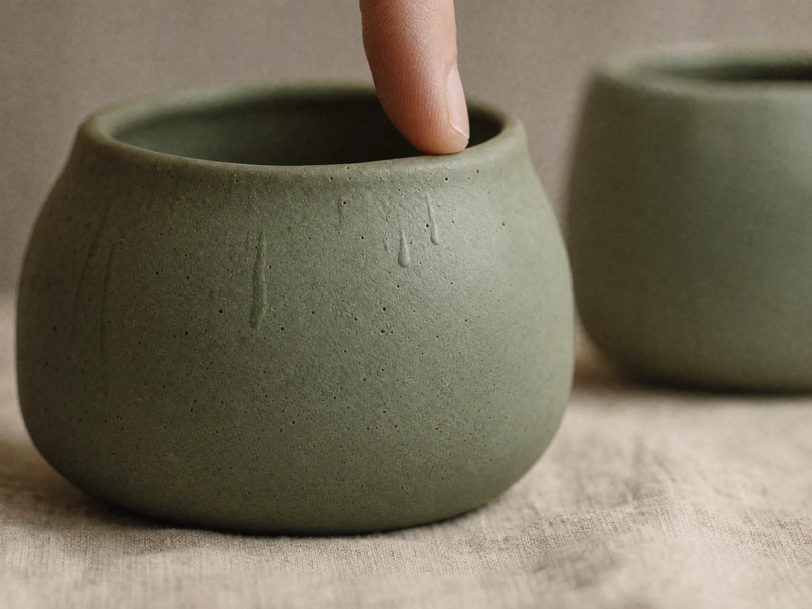Macro detail of a single sage-glazed ceramic vessel, matte iron-oxide rim catching window light